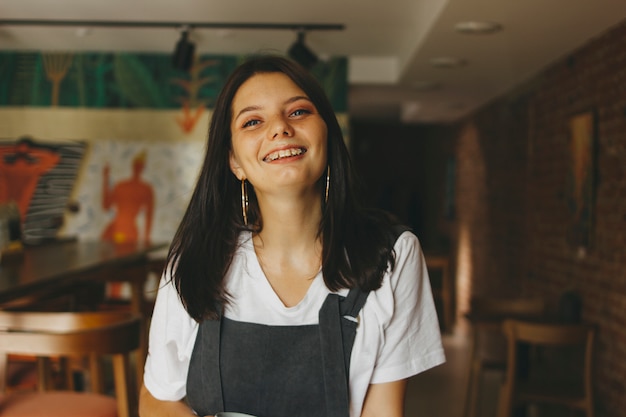 Portret van glimlachend charmant donkerbruin meisje met steunen op tanden in vrijetijdskleding in koffie