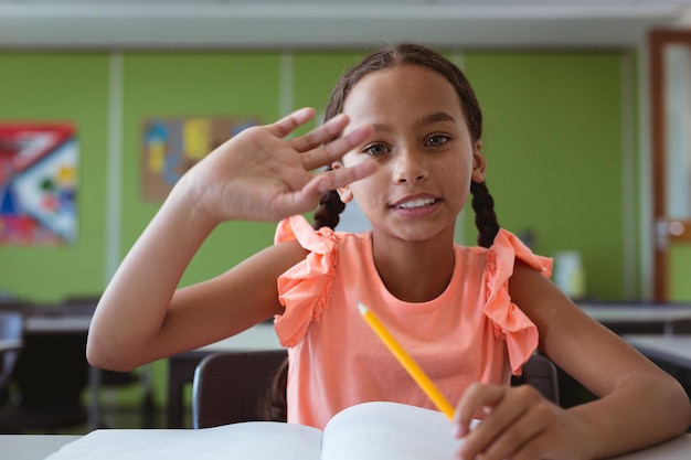 Portret van glimlachend gemengd ras schoolmeisje zittend aan een bureau in de klas die hand opsteekt