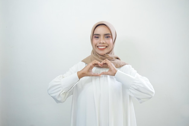 Portret van glimlachende Aziatische moslimvrouw die hartgebaar toont dat over witte achtergrond wordt geïsoleerd