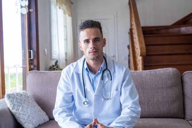 Foto portret van glimlachende blanke jonge mannelijke arts thuis in wit medisch uniform en stethoscoop zit op de bank kijk naar de camera gelukkige positieve man huisarts of arts poseert op de werkplekxa