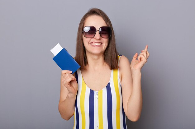 Portret van glimlachende hoopvolle vrouw die zwempak en zonnebril draagt, kruist de vingers en maakt de wens om naar het buitenland te reizen met paspoort en kaartjes die geïsoleerd op een grijze achtergrond staan