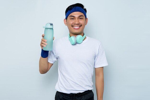 Portret van glimlachende jonge Aziatische sportieve man in blauwe hoofdband en witte tshirt met koptelefoon Holding waterfles geïsoleerd op witte achtergrond Workout sport concept
