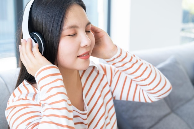 portret van glimlachende jonge aziatische vrouw met gesloten ogen die thuis muziek luistert in een draadloze hoofdtelefoon