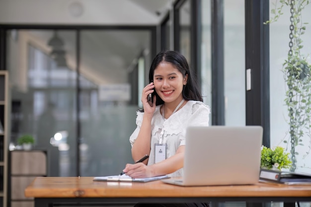 Portret van glimlachende jonge Aziatische vrouwelijke manager die op afstand werkt, papier vasthoudt en financieel document leest, op de mobiele telefoon praat, aan het bureau staat in het kantoor thuis