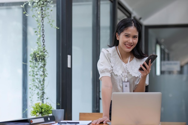 Portret van glimlachende jonge Aziatische vrouwelijke manager die op afstand werkt, papier vasthoudt en financieel document leest, op de mobiele telefoon praat, aan het bureau staat in het kantoor thuis