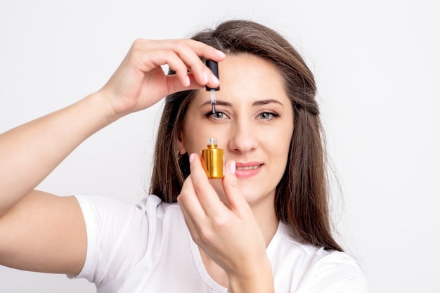 Portret van glimlachende manicuremeester houdt gouden nagellakfles over haar oog op witte muur