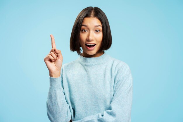 Foto portret van glimlachende mooie aziatische vrouw in casual kleding met vinger omhoog die een goed idee heeft en naar de camera kijkt, geïsoleerd op blauwe achtergrond.