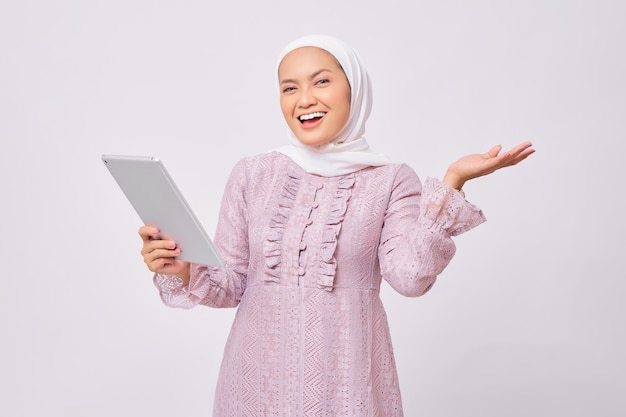 Portret van glimlachende mooie jonge Aziatische moslimvrouw die hijab en paarse jurk draagt die digitale tablet vasthoudt en hand presenteert op kopieerruimte geïsoleerd op witte studio achtergrond