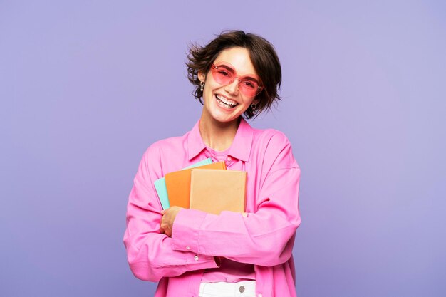 Foto portret van glimlachende mooie vrouwelijke student met boeken in een roze outfit die naar de camera kijkt