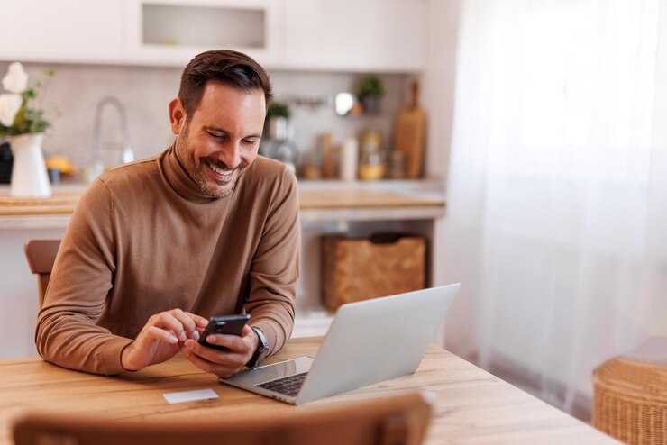 Foto portret van glimlachende volwassen man met creditcard met mobiele telefoon en laptop op eettafel thuis