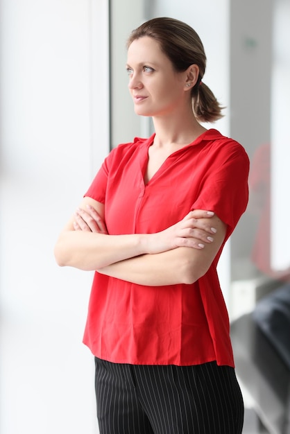 Portret van glimlachende vrouw in rode blouse gekruiste handen op de borst en uit het raam kijkend