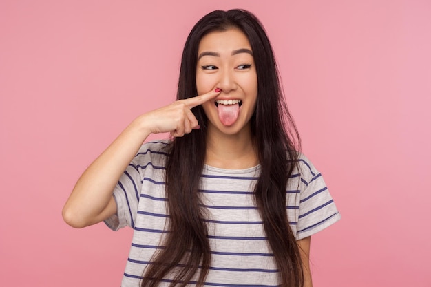 Portret van grappig komisch meisje met lang haar in gestreept t-shirt neus plukken, boogers trekken en tong tonen met humoristische dwaze uitdrukking. indoor studio-opname geïsoleerd op roze achtergrond