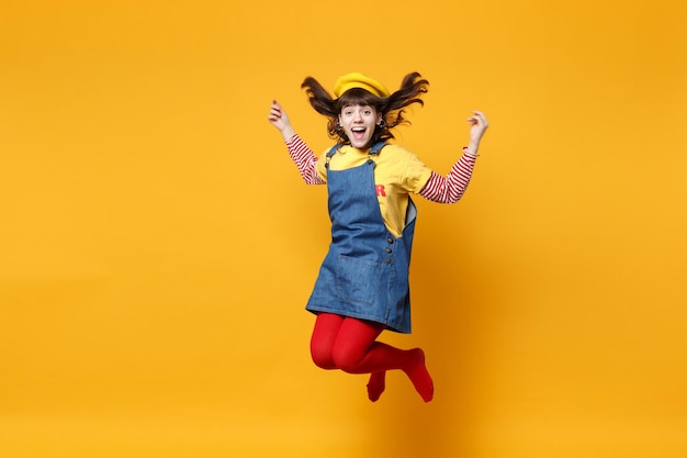 Portret van grappig meisje tiener in Franse baret en denim zomerjurk springen met fladderende haren geïsoleerd op gele muur achtergrond in studio. Mensen emoties, lifestyle concept. Bespotten kopie ruimte.