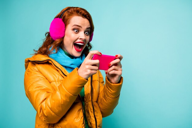 Portret van grappig verbaasd dame houden telefoon verslaafd videogames speler open mond opgewonden dragen trendy casual oorhoezen gele overjas sjaal.