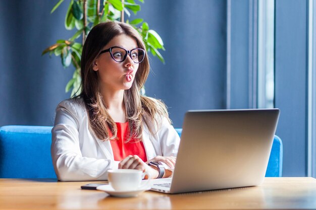 Portret van grappige gekke mooie brunette jonge vrouw in glazen zitten kijken naar haar laptop display met vis lippen en grote ogen grappig dwaas gezicht binnen studio shot café kantoor achtergrond