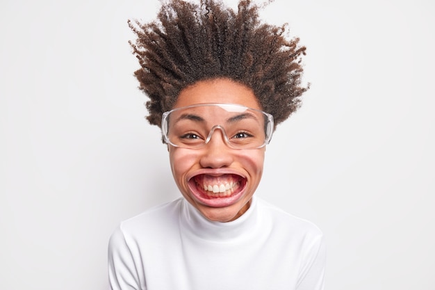 Portret van grappige positieve donkere vrouw glimlacht met tanden heeft grote mouh poses onder luchtdruk heeft haar opstaan gekleed in casual poloneck en grote transparante bril