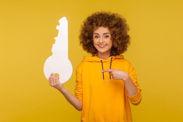 Portret van grappige positieve vrouw met krullend haar in stedelijke stijl hoodie wijzend op grote papieren sleutel en kijkend naar camera met brede glimlach, huisaankoop concept. studio-opname geïsoleerd op gele achtergrond