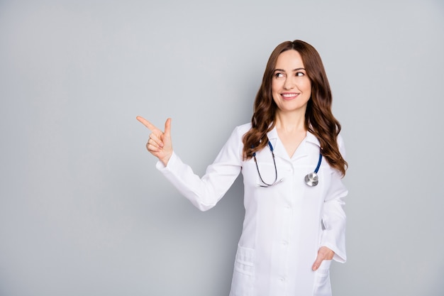 Portret van haar ze mooie aantrekkelijke vrolijke golvendharige dokter dragen jas phonendoscope stethoscoop tonen kopie ruimte blik idee geïsoleerd over grijze pastelkleur achtergrond