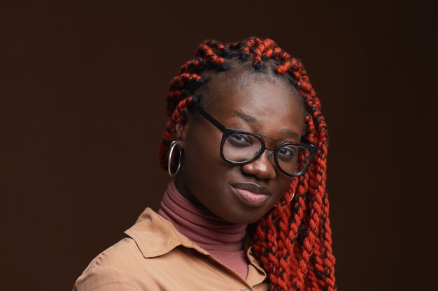 Portret van hedendaagse afro-amerikaanse vrouw met gevlochten haar camera kijken terwijl poseren tegen donkere bruine achtergrond in studio close-up, kopieer ruimte