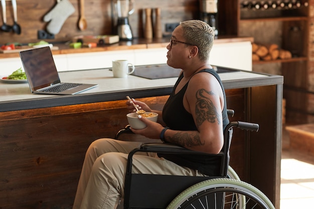 Portret van hedendaagse getatoeëerde vrouw in rolstoel die video's bekijkt via laptop thuis, kopieer ruimte