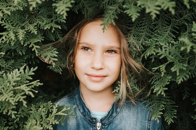 Portret van het gezicht van een schattig gelukkig Kaukasisch openhartig gezond achtjarig meisje omringd door takken en bladeren van groene plant thuja of cipres op de natuur buiten