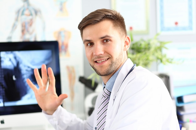 Portret van het glimlachen doc. Het stellen in het ziekenhuiskabinet. Glimlachende chirurg die medische laag draagt. X-ray foto van het lichaam op het display van de computer.