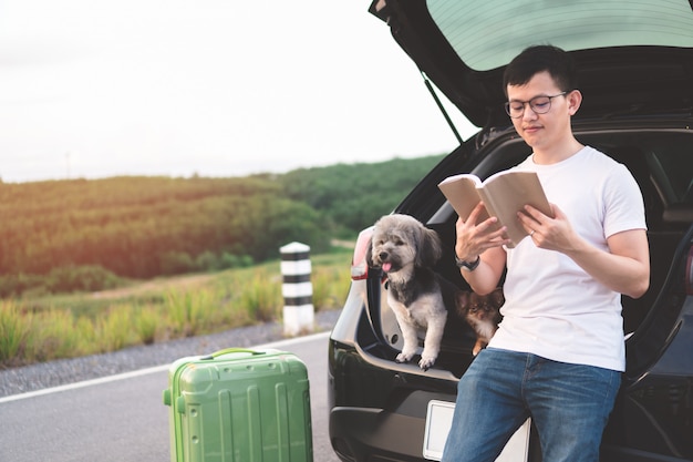 Portret van het jonge Aziatische boek van de mensenlezing terwijl het zitten in auto open boomstam met zijn honden.