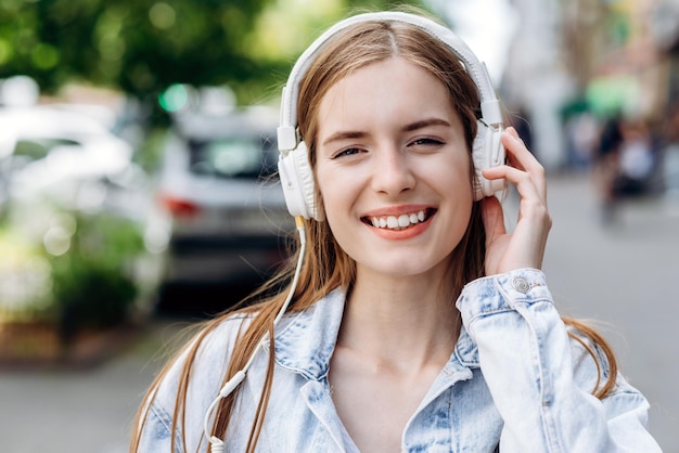 Portret van het jonge meisje dat muziek luistert in hoofdtelefoons stedelijke straatstijl Outdoor streetstyle hipster vrouw in hoofdtelefoons luistert naar muziek en glimlacht