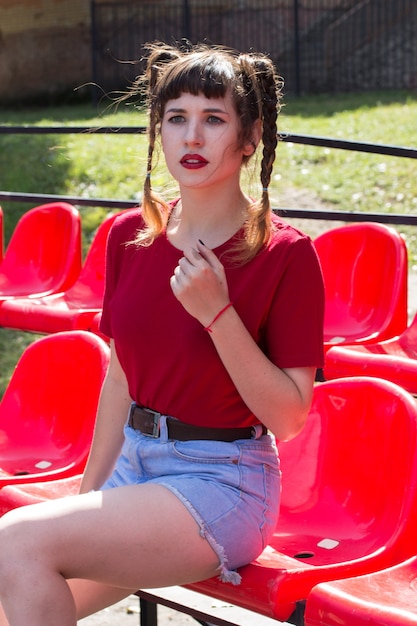 Portret van het jonge normale mooie model van het tennagermeisje in de zetels van een honkbalstadion dat rode t-shirt draagt