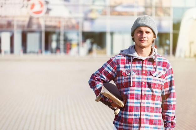 Portret van hipster man gekleed in cheked shirt en pet met skateboard voor modern gebouw