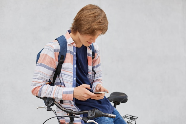 Portret van hipster man met trendy kapsel dragen shirt en spijkerbroek rugzak staande houden in de buurt van zijn fiets met behulp van slimme telefoon