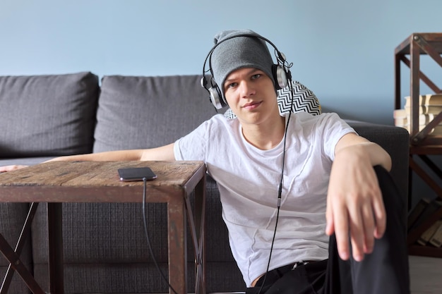 Portret van hipster tiener in hoed met koptelefoon zittend op de vloer thuis. Trendy knappe jongen die naar de camera kijkt. Jeugd, levensstijl en vrije tijd