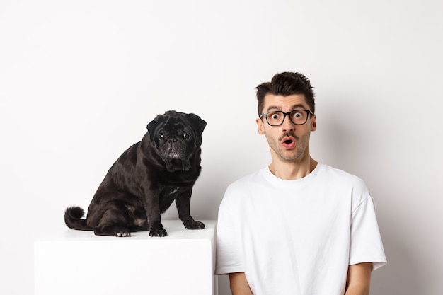 Portret van hondeneigenaar en kleine schattige mops die verbaasd en verbaasd naar de camera staren, staande op een witte achtergrond