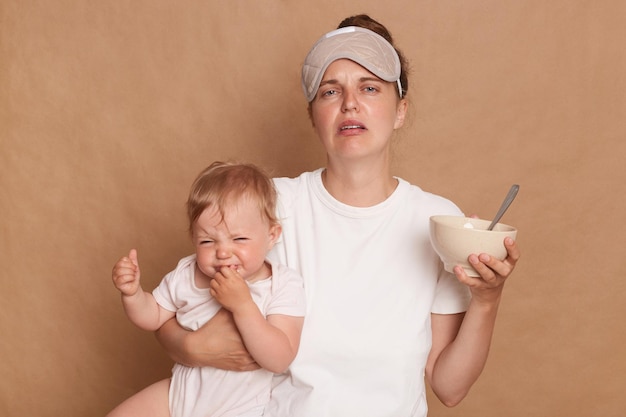 Portret van huilende, verdrietige, gestreste blanke vrouw, gekleed in een wit T-shirt en slaapmasker, geïsoleerd over een bruine achtergrond, babydochter en bord met voedsel vast te houden dat kind moet voeden