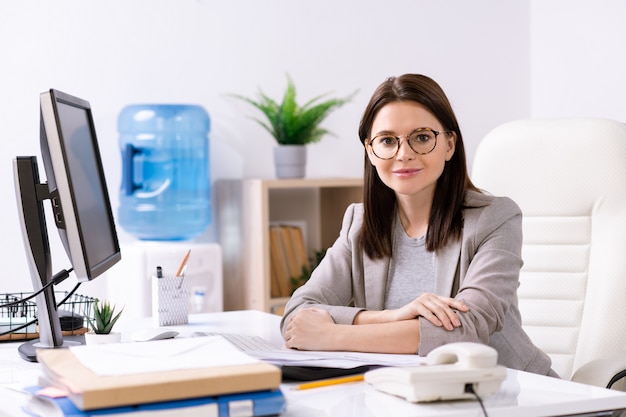 Portret van inhoud aantrekkelijke jonge officemanager in jasje zit aan bureau met computer, papieren en telefoon