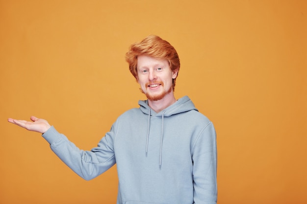 Portret van inhoud jonge blogger met rode baard in hoodie die opzij wijst terwijl hij advertenties op oranje doet