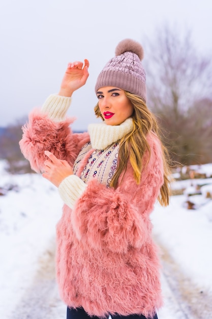 Portret van jong blond model dat roze bontjasje, de winterlaarzen en een purpere hoed in de sneeuw draagt. Levensstijl