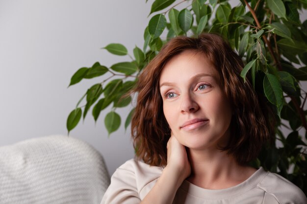 Portret van jong Kaukasisch donkerbruin meisje met krullen in een beige kleding op een achtergrond van een witte muur en groene bladeren van boom