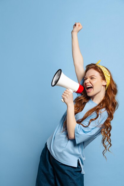 Portret van jong mooi roodharig meisje schreeuwend in megafoon geïsoleerd over blauwe studio achtergrond