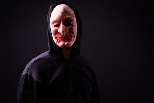 portret van jonge Aziatische man met hoodie en horror masker op zwart