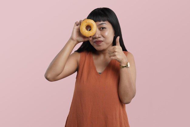 Portret van jonge Aziatische vrouw met donuts met duim omhoog vinger, geïsoleerd over roze background