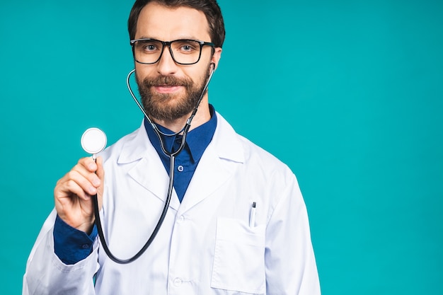 Portret van jonge, bebaarde dokter man met stethoscoop over nek in medische jas staande tegen geïsoleerde blauwe achtergrond.