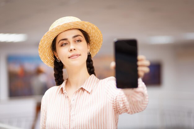 Portret van jonge blanke mooie vrouw in strohoed is toont smartphone met zwart scherm Op de achtergrond is kunst tentoonstelling zaal van galerie met schilderijen Concept van cultureel onderwijs