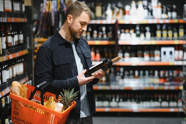 Portret van jonge blije positieve mannelijke klant die wijn in supermarkt selecteert