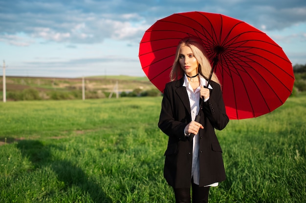 Portret van jonge blonde mooi meisje in zwart-wit pak, met rode paraplu tegen de achtergrond van bewolkte hemel en groen veld.