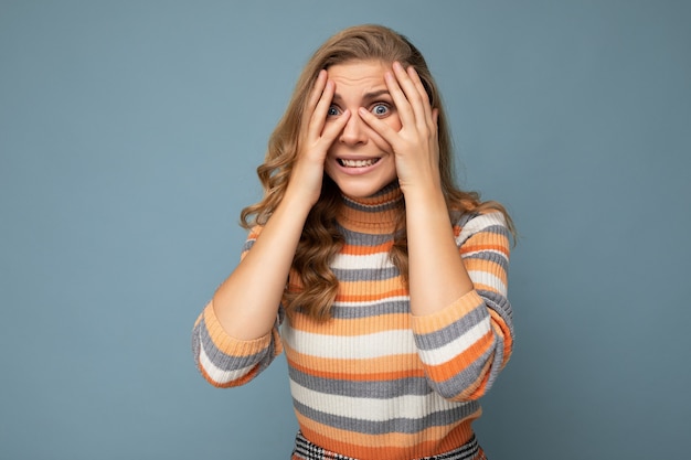 Portret van jonge blonde mooie krullende vrouw met oprechte emoties dragen gestreepte trui geïsoleerd op blauwe achtergrond met lege ruimte en bang gezichtsuitdrukkingen verbergen.