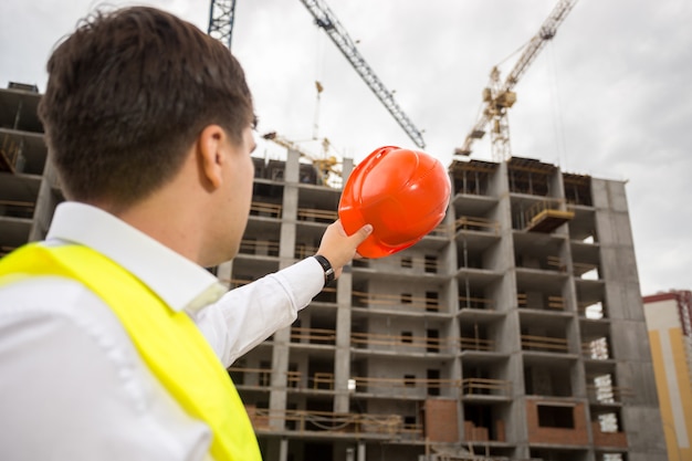 Portret van jonge bouwingenieur wijzend op gebouw in aanbouw met rode veiligheidshelm