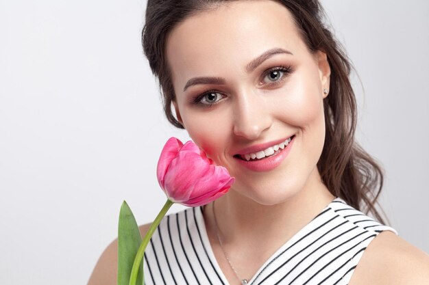 Portret van jonge brunette mooie vrouw met make-up in gestreepte jurk, permanent en roze tulp vasthouden en camera kijken met brede glimlach. indoor studio opname, geïsoleerd op lichtgrijze achtergrond.