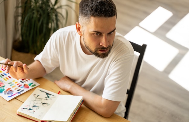 Portret van jonge donkerharige geconcentreerde blanke blanke mannelijke artiest met een baard in een wit t-shirt in studio verlicht door zonlicht. zittend in een studio in houten tafel. in een hand met kleurpotlood