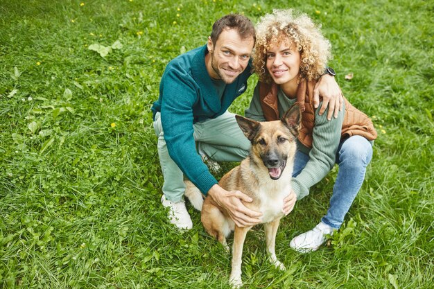 Portret van jonge gelukkige paar zittend op groen gras met Duitse herder en glimlachend in de camera